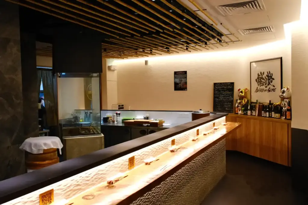 An intimate standing sushi counter with warm under-counter lighting, bamboo ceiling accents, and Japanese calligraphy artwork at Sushi to Sumi in Raffles Place.