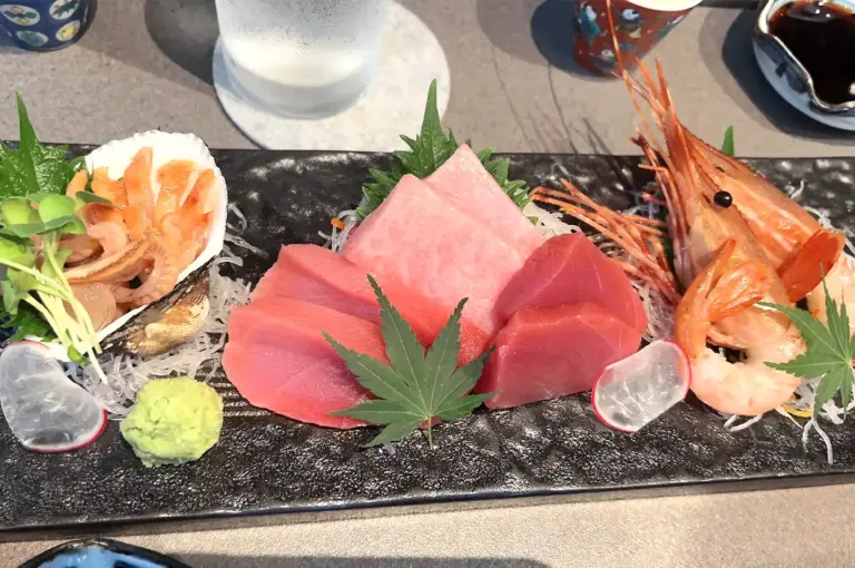 Assorted sashimi platter featuring fresh tuna cuts, sweet prawns, and clams served with wasabi and radish on a dark stone plate.