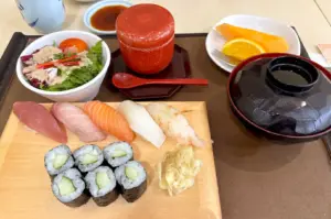 A fresh sushi platter featuring assorted nigiri, cucumber maki rolls, a side salad, miso soup, and fresh fruit.