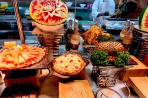 A buffet table featuring a intricately carved watermelon centerpiece, sliced poultry cold cuts, a freshly baked pizza, roasted chicken, and decorative spice bottles.