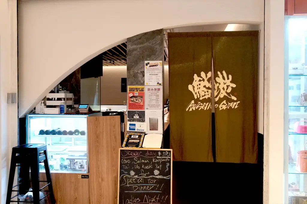 The entrance of Sushi To Sumi featuring a dark noren curtain with traditional Japanese lettering, a standing chalkboard menu showcasing special skewer options like otoro and salmon, and a beverage display fridge.