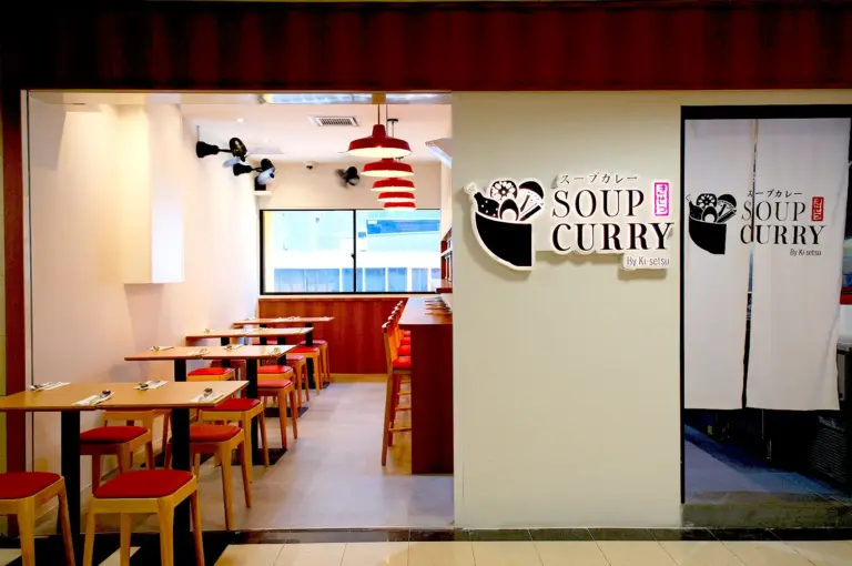 A welcoming storefront of Soup Curry by Ki-setsu in Singapore, featuring a modern Japanese sign, red pendant lamps, and a cozy dining room with wooden tables and chairs.