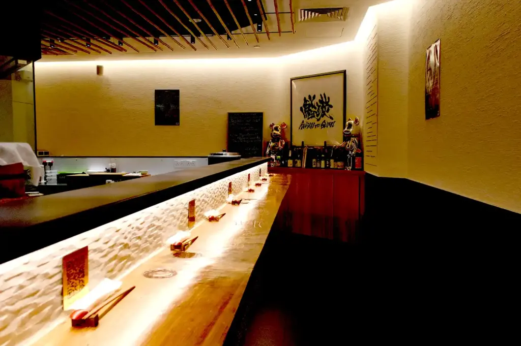 The intimate, warmly lit interior of Sushi To Sumi featuring a clean wooden counter bar with individual place settings, textured white walls, and Japanese artwork.