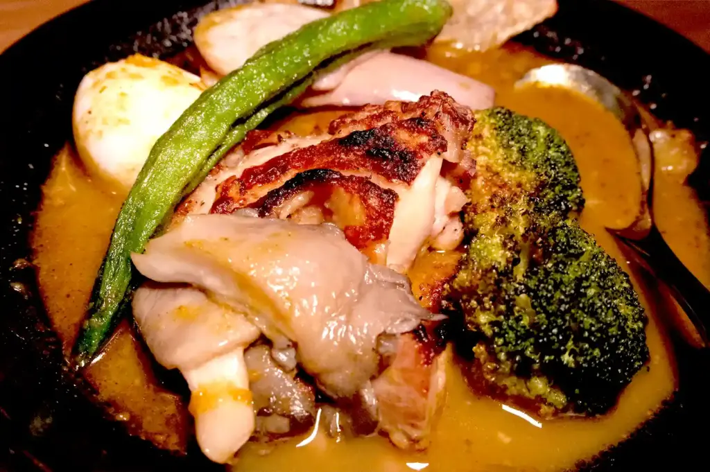 A close-up of a rich, aromatic Hokkaido soup curry loaded with tender chicken, a soft-boiled egg, fresh okra, and charred broccoli in a black bowl.