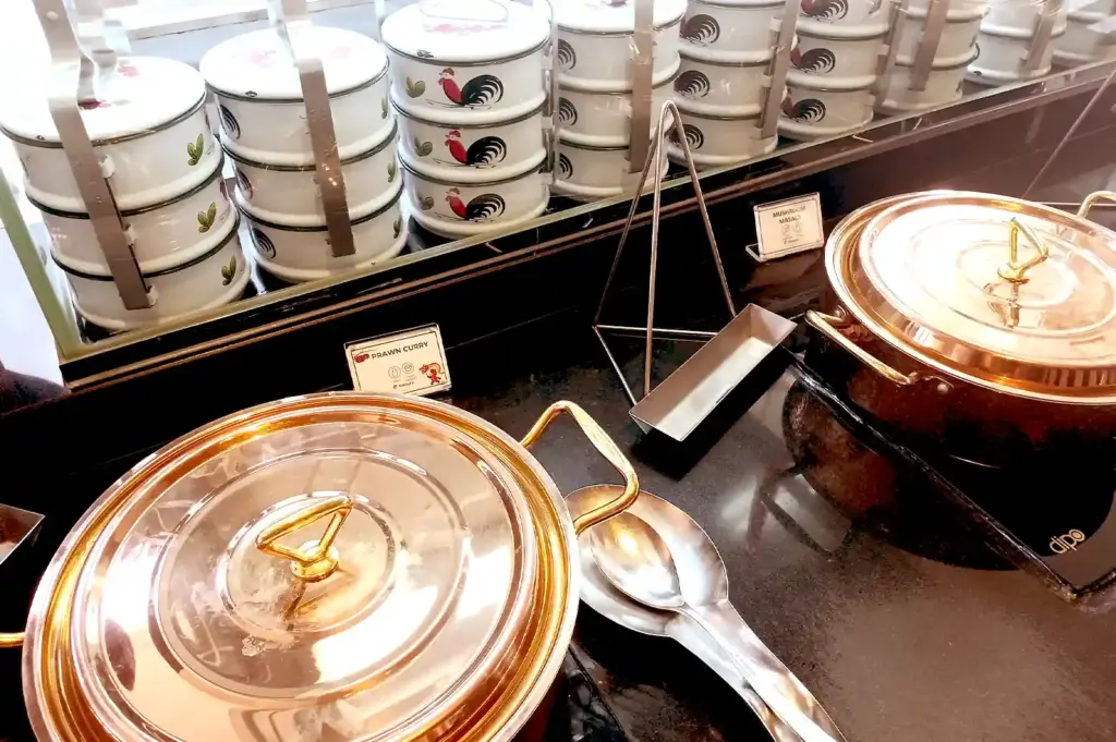 Traditional rooster-patterned tingkat carriers stacked beside polished copper pots containing dishes like prawn curry and mushroom masala