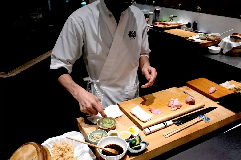 A sushi chef in traditional white attire preparing ingredients at the wooden counter, surrounded by fresh cuts of fish, bowls of wasabi, and condiments.