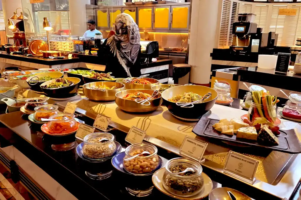 A buffet layout showcasing bowls of salads, cheeses, assorted nuts, dried fruits, and sliced vegetables under warm overhead lighting.