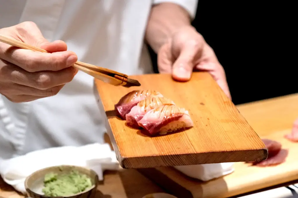 Close-up of a chef's hands using chopsticks to precisely brush a sweet glaze over fresh pieces of nigiri sushi served on a wooden board.
