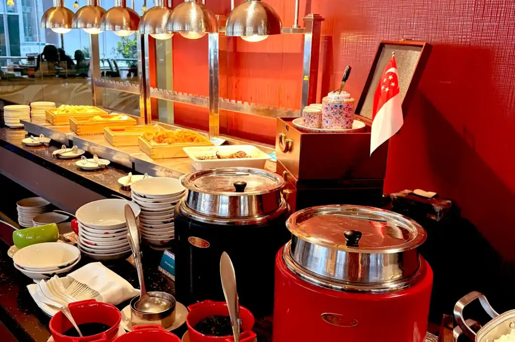 A buffet counter lined with warm food warmers, soup pots, stacked plates, and small bowls accompanied by miniature Singapore flags.