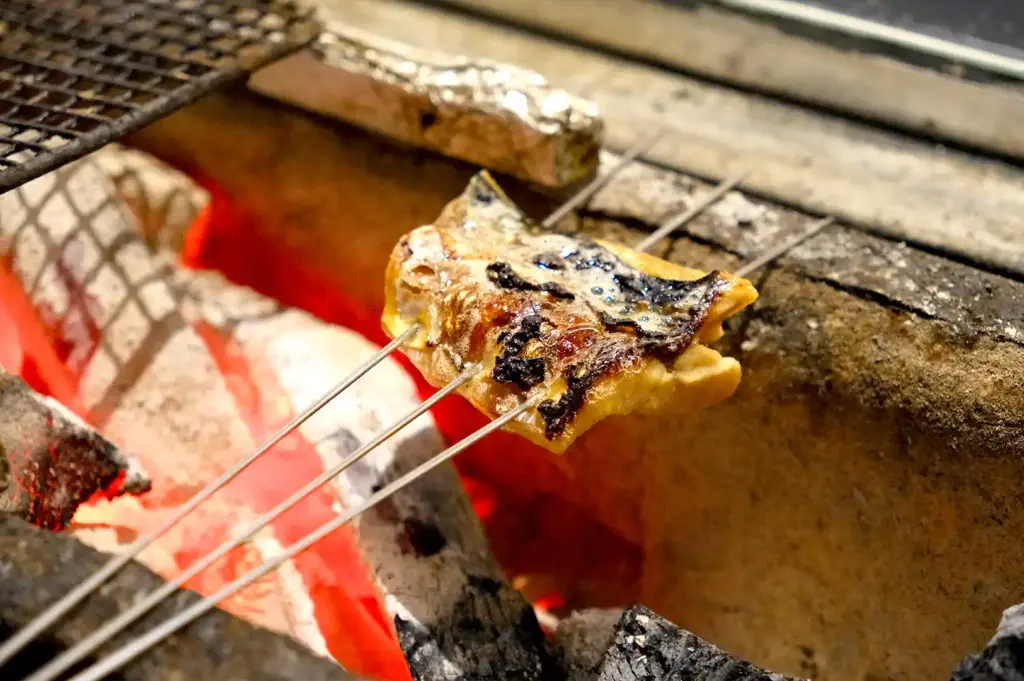 A piece of fish grilling over glowing charcoal in a traditional Japanese robata grill, highlighting the restaurant's sumi (charcoal) cooking philosophy.