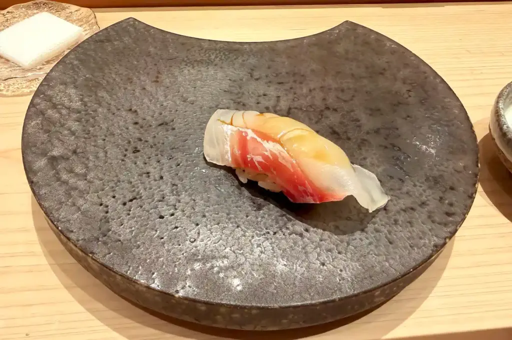 A single, masterfully crafted piece of nigiri sushi resting on an artistic dark stone plate at Sushi Sato.