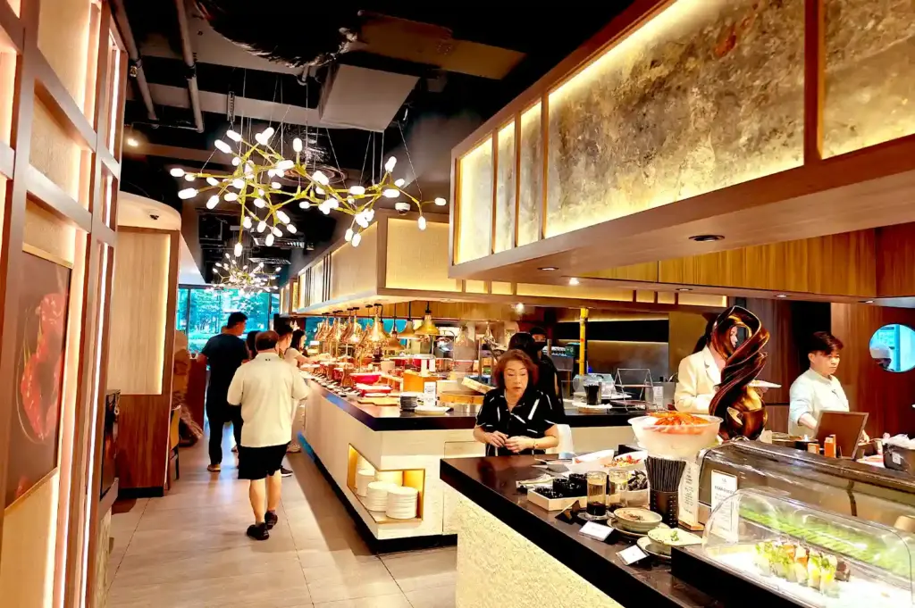Diners walking through a modern, warmly lit restaurant interior with elegant overhead light fixtures and a sushi and seafood counter.