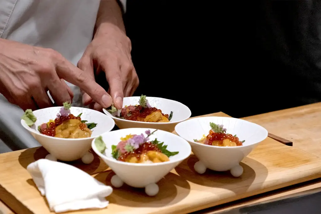 A chef carefully garnishing small bowls of rice topped with sea urchin (uni) and salmon roe (ikura) served in elegant white ceramic bowls.