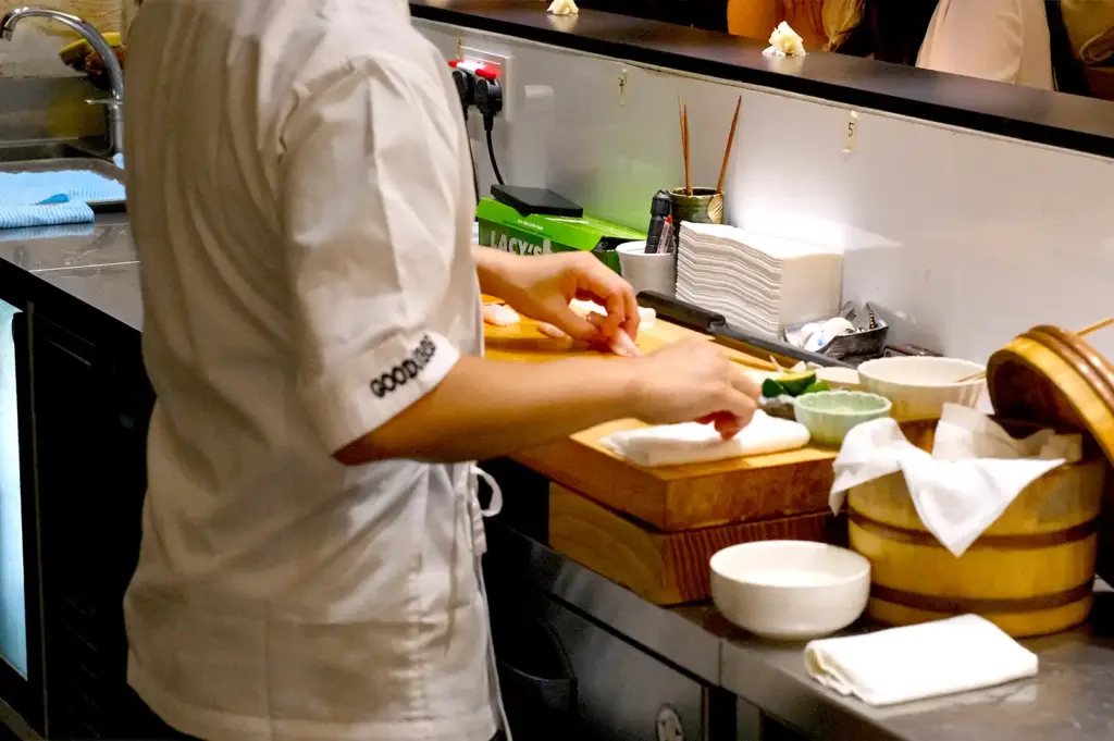 A sushi chef seen from behind while meticulously slicing and preparing fresh seafood on a traditional wooden cutting board at the preparation station.