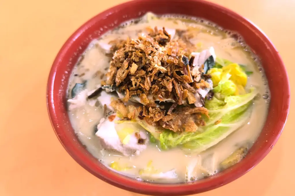 Close-up overhead shot of a steaming bowl of milky fish soup topped with generous fried shallots, fresh sliced fish, and cabbage in a red bowl.