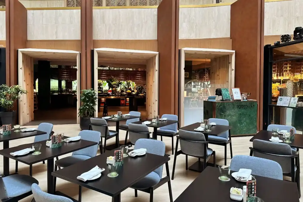 High-ceiling dining atrium at Peppermint restaurant featuring wooden arches, dark dining tables with grey armchairs, and an open buffet station in the background.