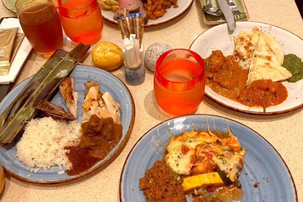 Dining table spread with plates of Asian, Mediterranean, and Indian buffet dishes alongside fresh drinks at Carousel Buffet.