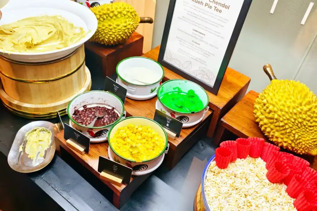 Chendol dessert station at Peppermint buffet displaying durian pengat in a wooden tub, red beans, sweet corn, green jelly noodles, and Kueh Pie Tee shells.